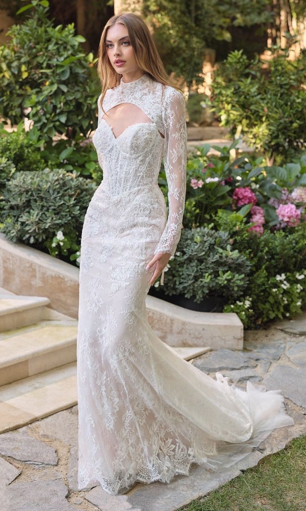 A woman in the Andrea and Leo WL071 long wedding dress, featuring fitted beaded lace, a strapless sweetheart neckline, and sheer overlay, stands outdoors by garden steps and flowers.